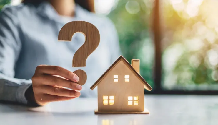 a hand holding-forward a wooden carved question mark and a similar looking home beside it